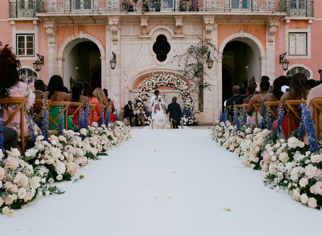 Mo & Toba kneeling at a floral arch at the end of a white aisle lined with roses, Lisbon